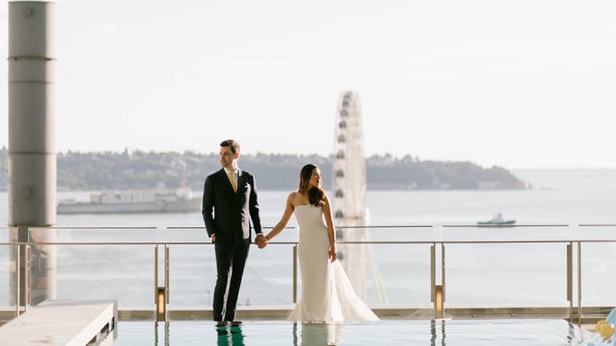 Bride and groom hold hands on sunny outdoor swimming pool deck and glass balcony overlooking water