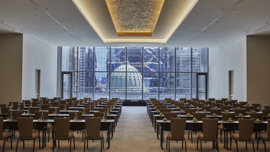 A large meeting room with rows of tables and chairs facing a podium with a wall of windows behind it.