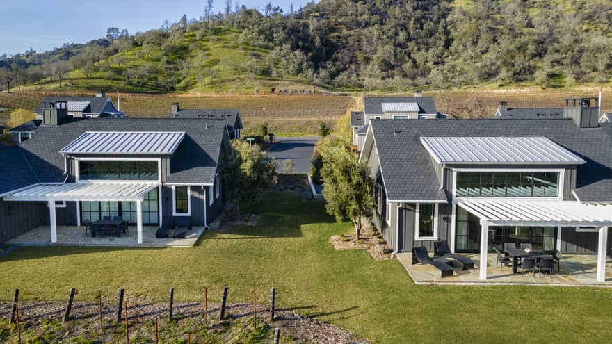 Aerial view of luxury resort gray villas with patios with pergolas, surrounded by grassy lawns, vineyards and hillside scenery