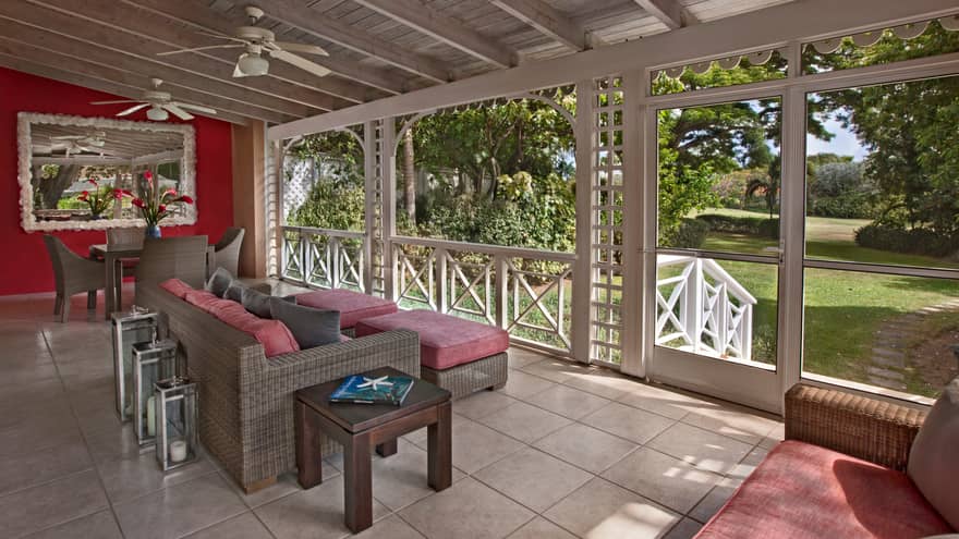 Wicker table, sofas with red cushions under wood beam roof, screened-in veranda with white trim