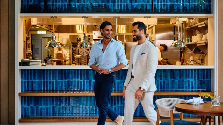 Two guests ?�one wearing a white suit and white t-shirt and the other wearing a light-blue dress shirt and dark-blue dress pants ?�walk by an open kitchen in a restaurant dining room
