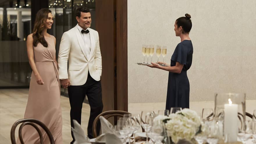 Hotel staff holding tray with Champagne glasses greets couple wearing formal attire in banquet room
