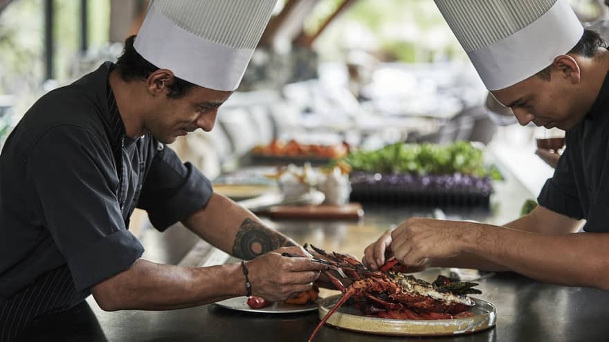 Two chefs in uniform assemble grilled lobster entree on long kitchen counter 