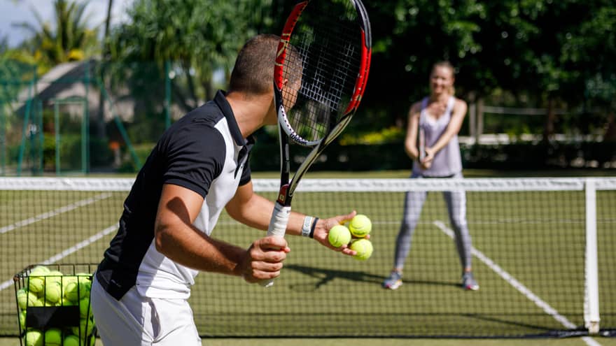 Couple playing tennis on court, man prepares to serve tennis balls to woman ready to swing