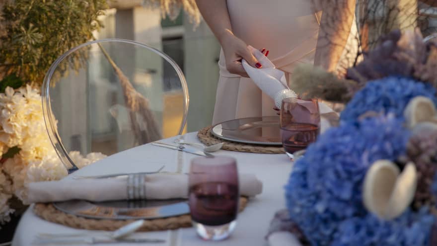 Woman sets rolled napkin on outdoor dining table beside tall floral planter