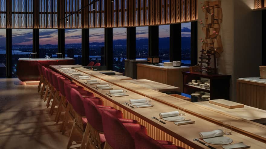 Ten place settings at a long wooden table with warm underlighting, a curved bank of windows offering a dramatic sunset view.