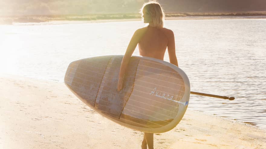 Silhouette of woman walking with large paddle board across beach at sunrise 