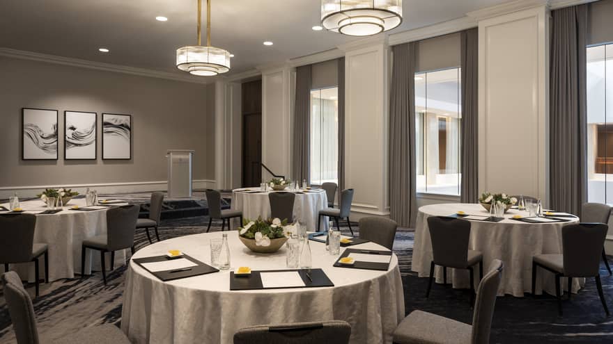A room with large round tables covered in cloth. A menu is placed at each seat, while a flower arrangement is at the center of each table.