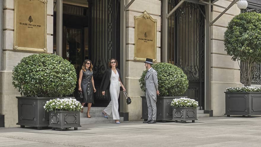 Four Seasons luxury hotel entrance with two guests walking out, doorman, glass canopies and large potted plants
