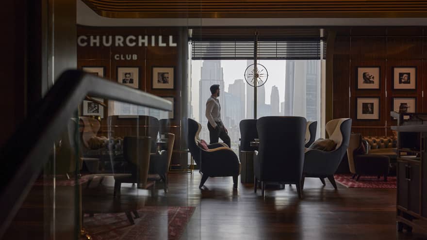 Man stands by a floor-to-ceiling window looking out on the city skyline in a dimly lit cigar bar filled with club chairs and leather sofas