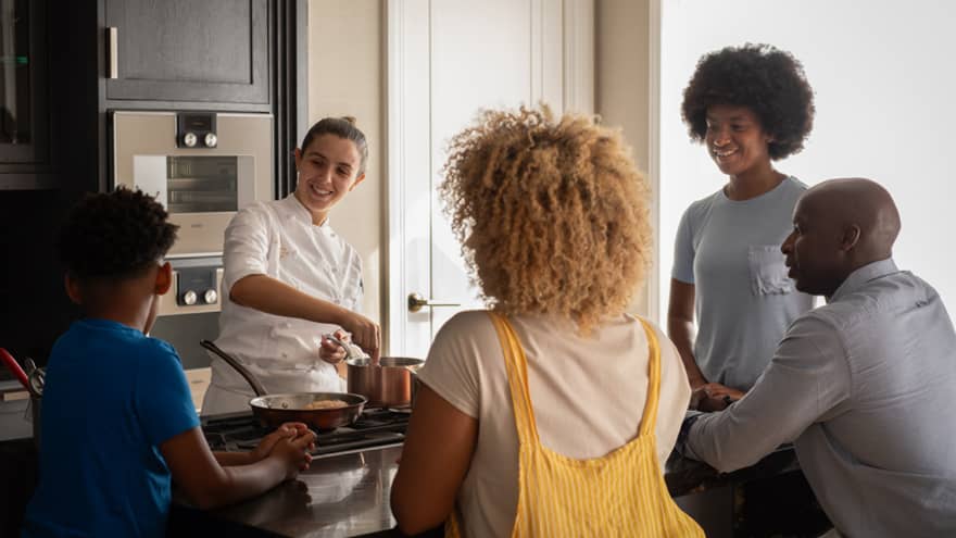 A family of four gathers around a kitchen-island cooktop watching a smiling chef prepare food in copper cookware. 
