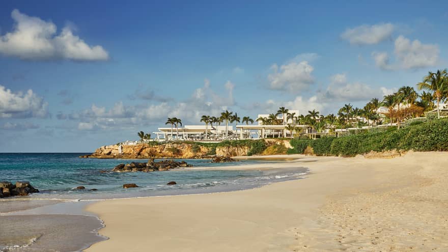 View across white sand beach to modern white event venue on rocks over sea 