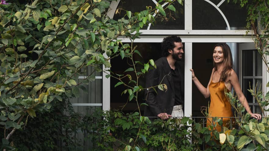 Two people stand on a vine-covered balcony surrounded by foliage.