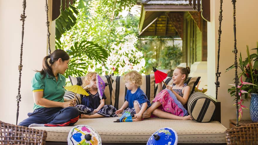 Hotel staff relaxes with children, hold colourful paper umbrellas on large hanging patio bed 