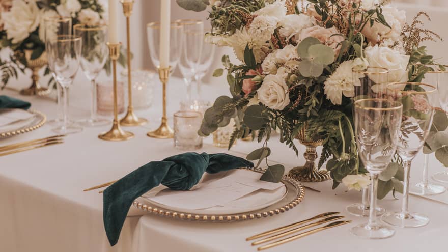 A beautifully arranged table adorned with gold cutlery, white roses and dark green knotted napkins, setting the vibe for a perfect wedding dinner