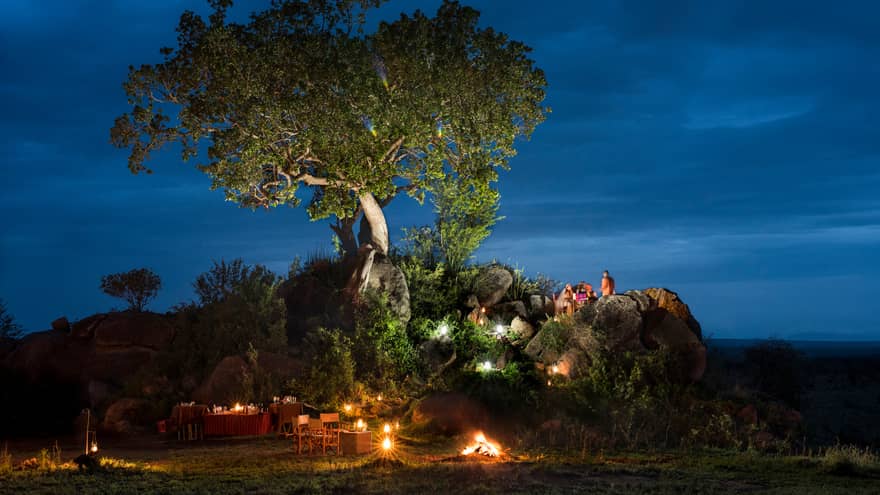 Bride and groom sit on large rock under tree, over candle-lit dining tables at night