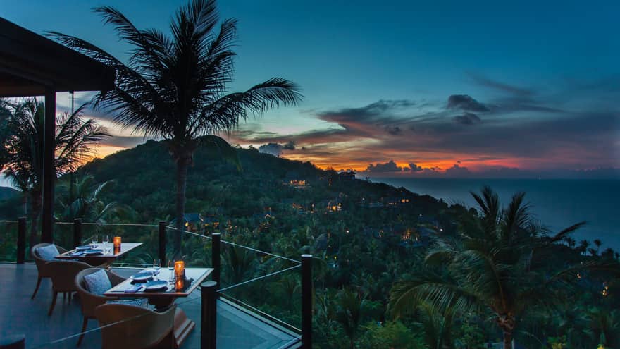 Corner of extended balcony with dining tables and chairs at dusk, expanse of forest, palm trees