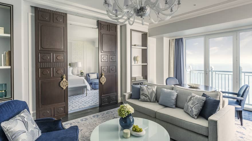 Sea-view living room, with grey sofa, blue pillows and accent chairs, chandelier, dark wooden doors open to bedroom
