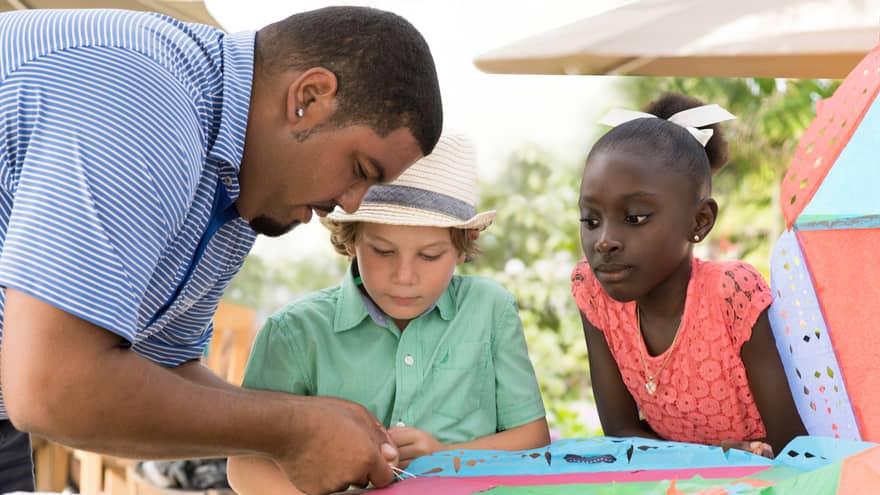 Man ties string onto colourful paper kite at table as two children watch
