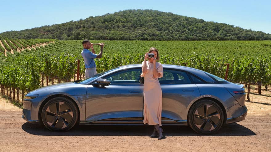 Person wearing long light-pink dress stands leaning against a grey car taking a picture of the surrounding vineyards with their camera while another person wearing a blue shirt and white pants stand on the other side of the car