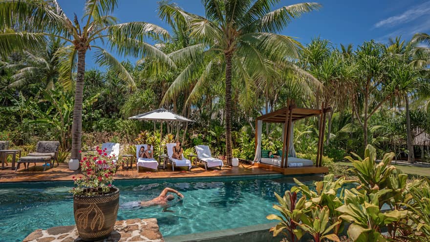 A man swims in an outdoor pool surrounded by palm trees, adjacent dek has white lounge chairs and umbrellas