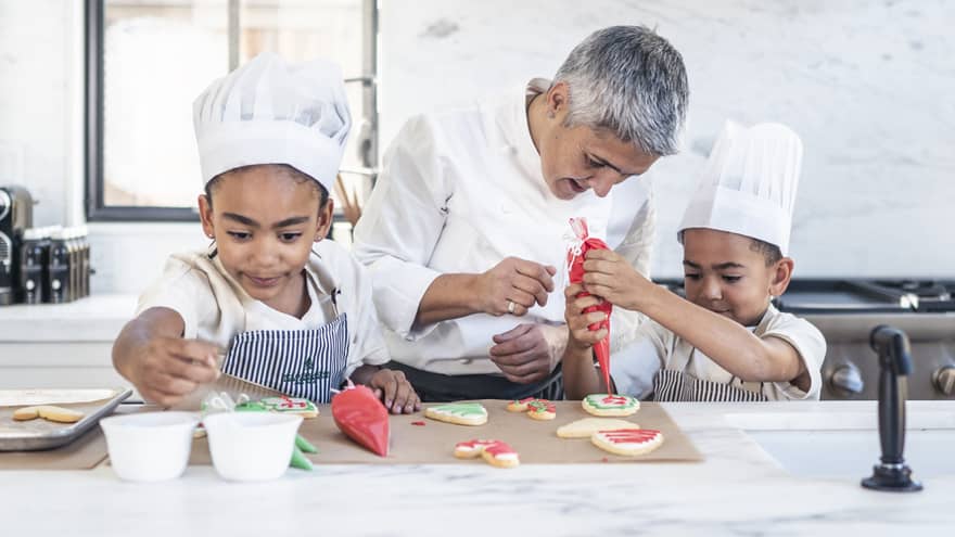 Two children in chef’s hats and aprons in a stark, white kitchen; a chef watches closely as one pipes icing onto a cookie.