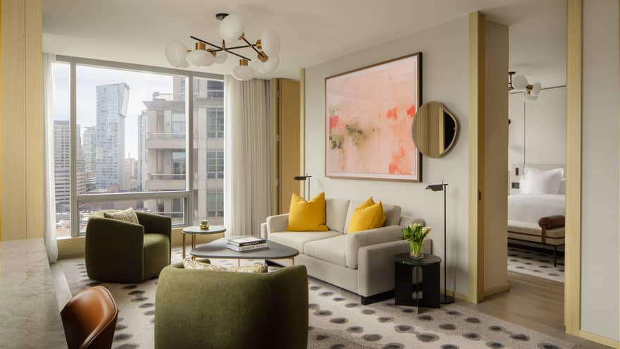 Living area with a beige sofa, yellow accent pillows, green armchairs, abstract artwork, and large windows with city views, connected to a bedroom
