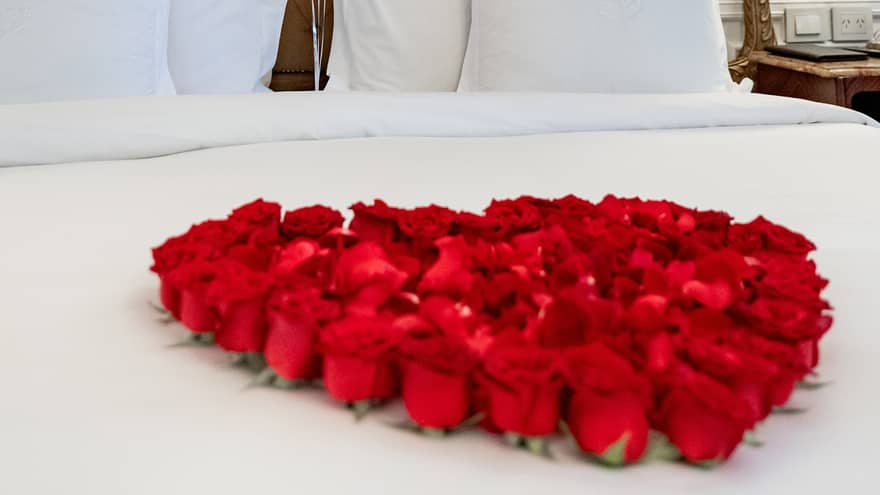 Red roses are placed on a white bed in the shape of a heart with three red heart-shaped balloons floating near the headboard