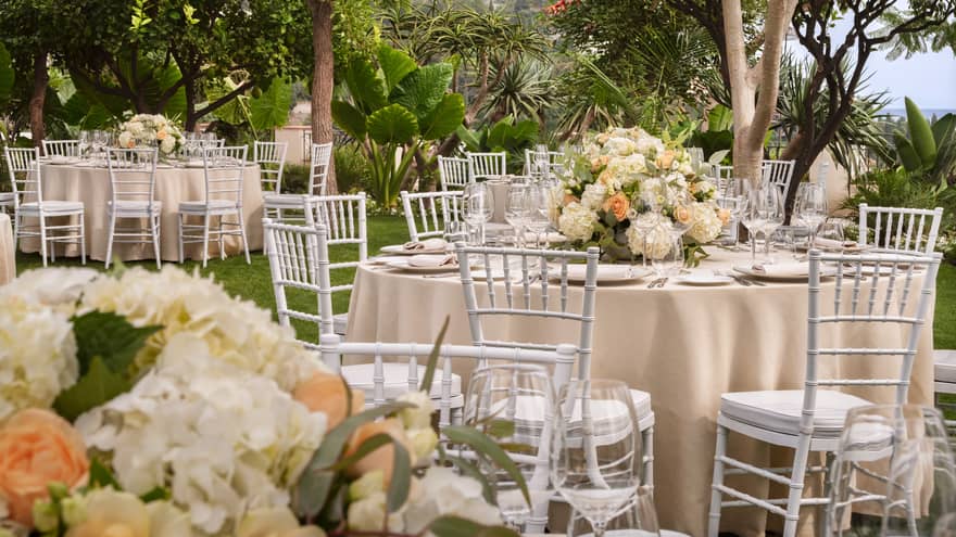 Outdoor wedding reception featuring white chairs and tables adorned with peach and ivory flowers in a garden setting