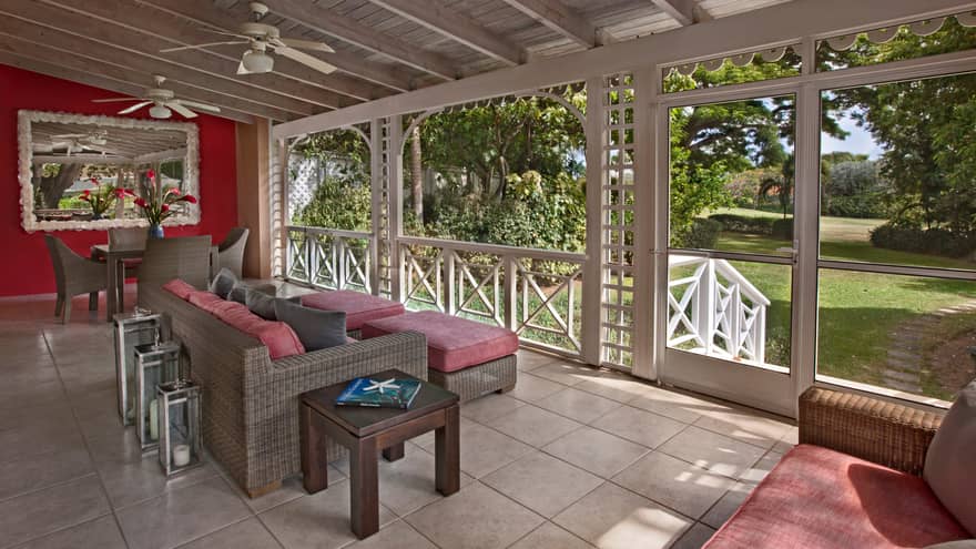 Wicker table, sofas with red cushions under wood beam roof, screened-in veranda with white trim