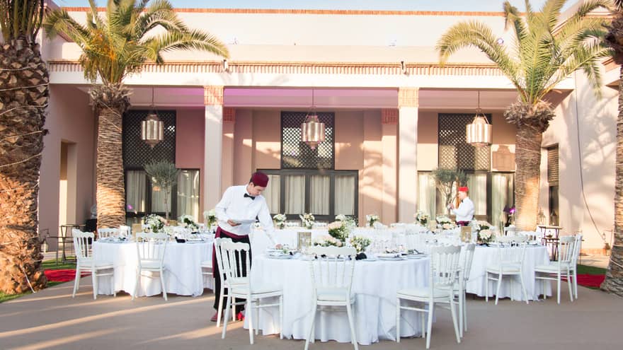 Hotel staff set white wedding banquet tables on sunny outdoor patio by pink building