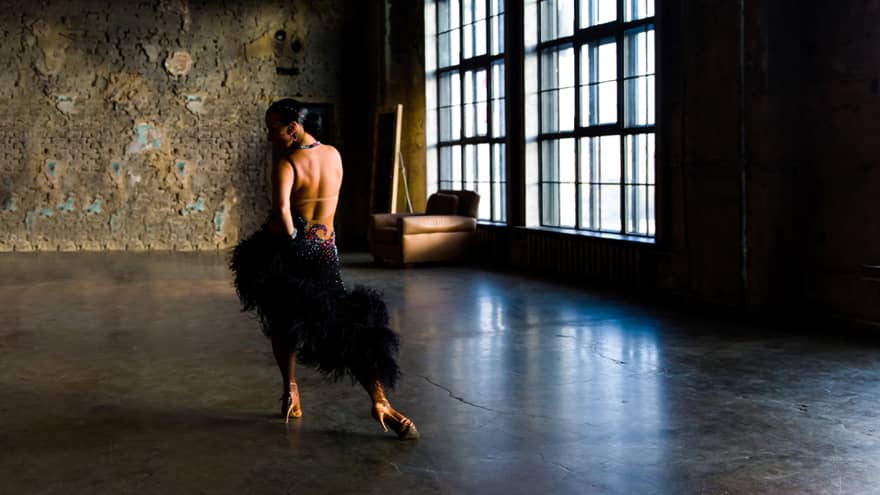 In an unlit room with large windows, a salsa dancer in an elaborate costume poses, hand on hip, opposite a brick wall. 