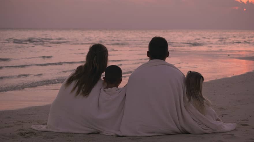 Back of family wrapped in white towels sitting on white sand beach at sunset