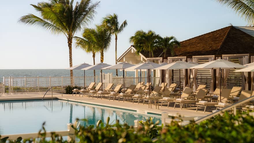 Outdoor pool lined with white lounge chairs an umbrellas with the beach in the background