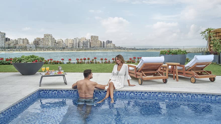 Man stands in pool, woman sits at pool's edge, sea in the background