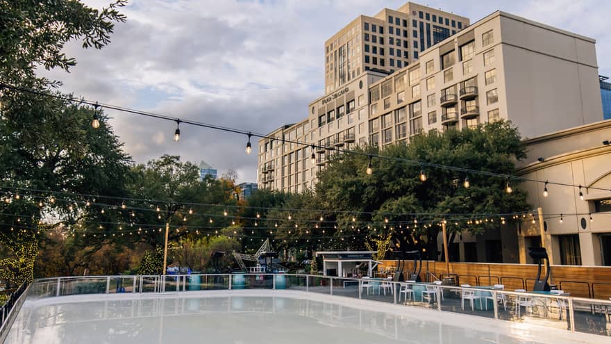 View of the Hotel overlooking an outdoor skating rink surrounded by trees and tables and lit by strings of Edison lights.