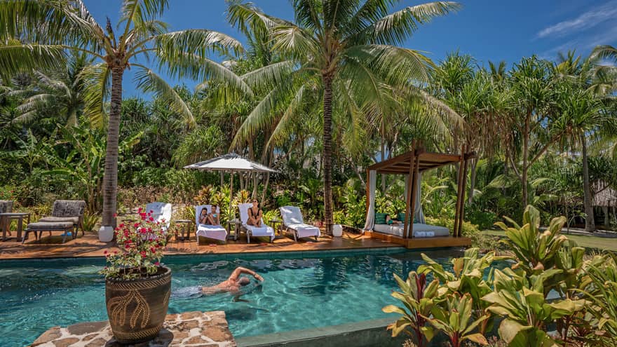 A man swims in an outdoor pool surrounded by palm trees, adjacent dek has white lounge chairs and umbrellas