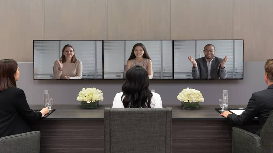Several people in business clothes sit in front of a series of screens in a virtual meeting.