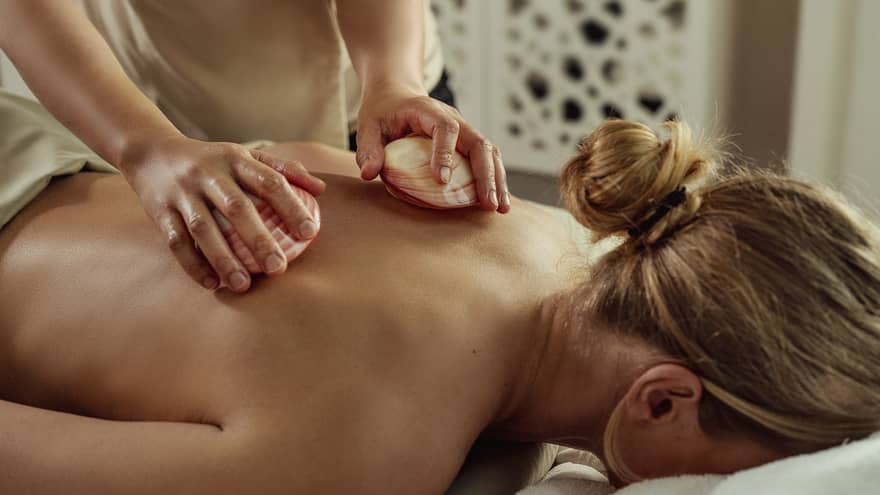 Guest lies face down on a massage table as a massage therapist rubs lava shells along their bare back