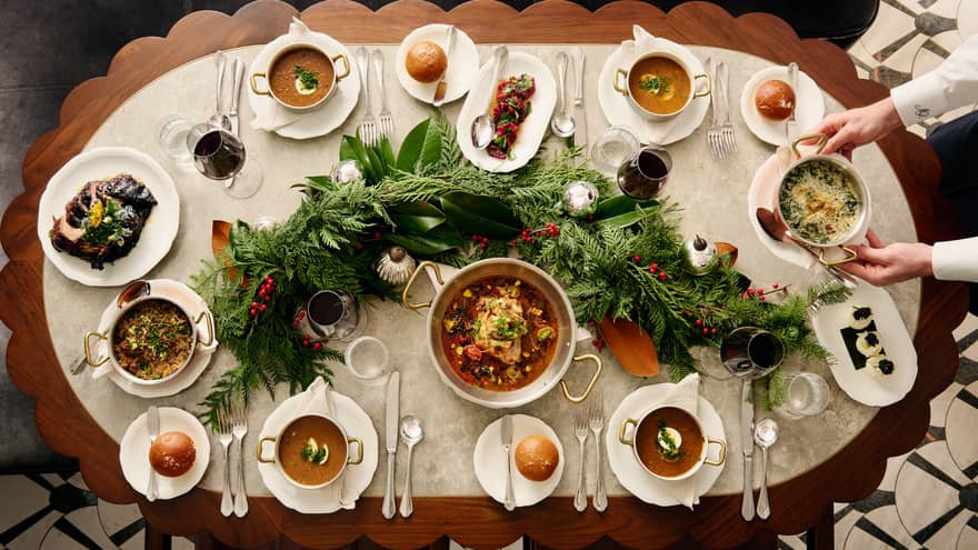 View from above of hands placing a dish on a table laden with soups, stews, bread and wine and adorned with festive greenery.
