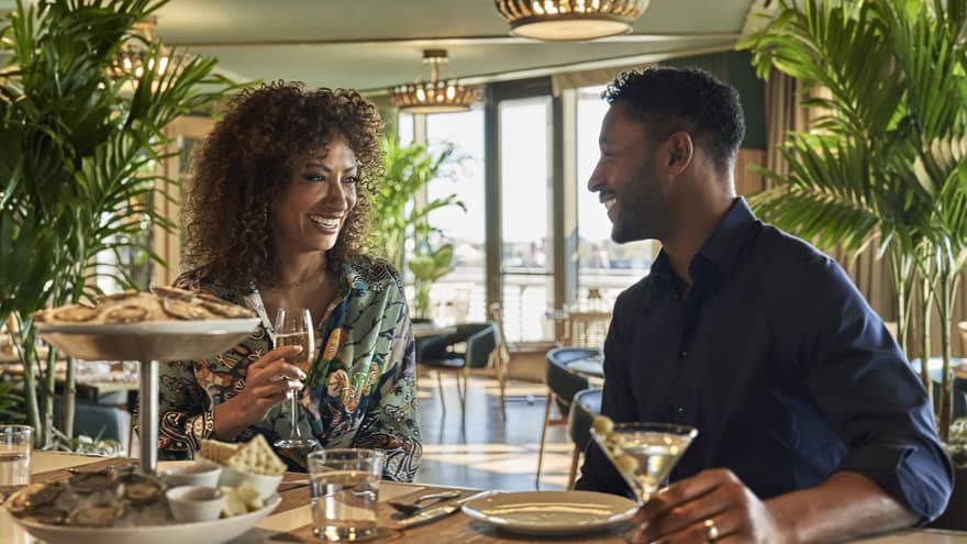 A man and woman eating oysters at an oyster bar with plants behind them.