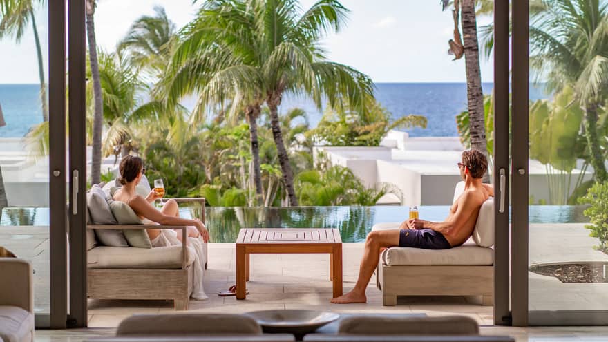 Couple relaxing on lounge chairs and enjoying drinks on a luxury resort private terrace overlooking tropical palm trees, infinity pool and the ocean