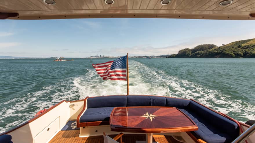 The back of a yacht, with a red table and an American flag, moving along the water.