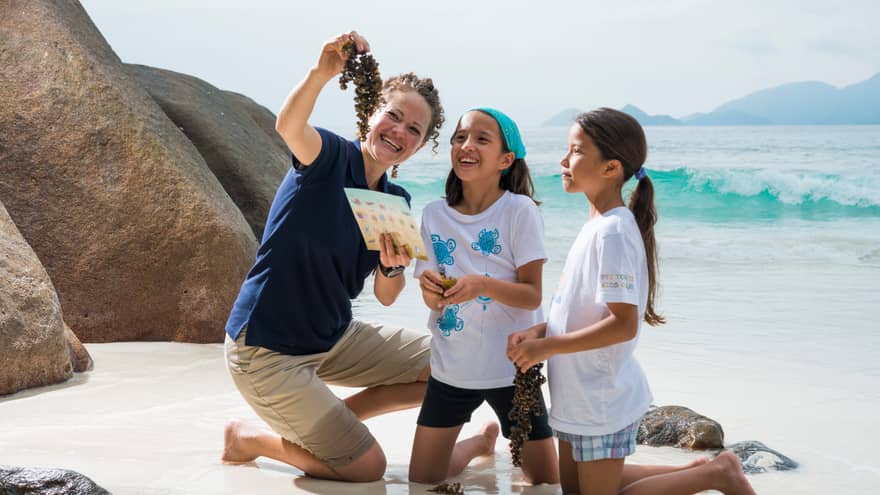 Woman kneels on white sand beach beside boulder, holds up seaweed to show two girls