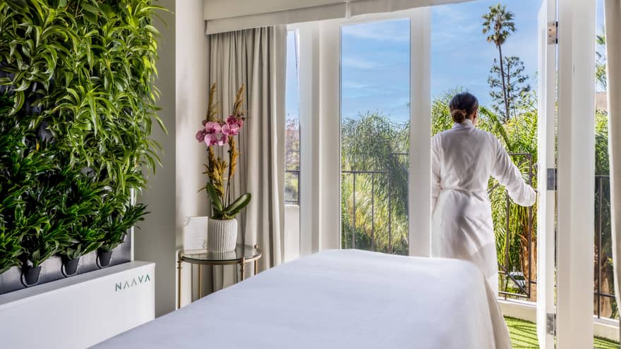 A woman walking outside from a massage room with large windowed doors and plants on one wall.