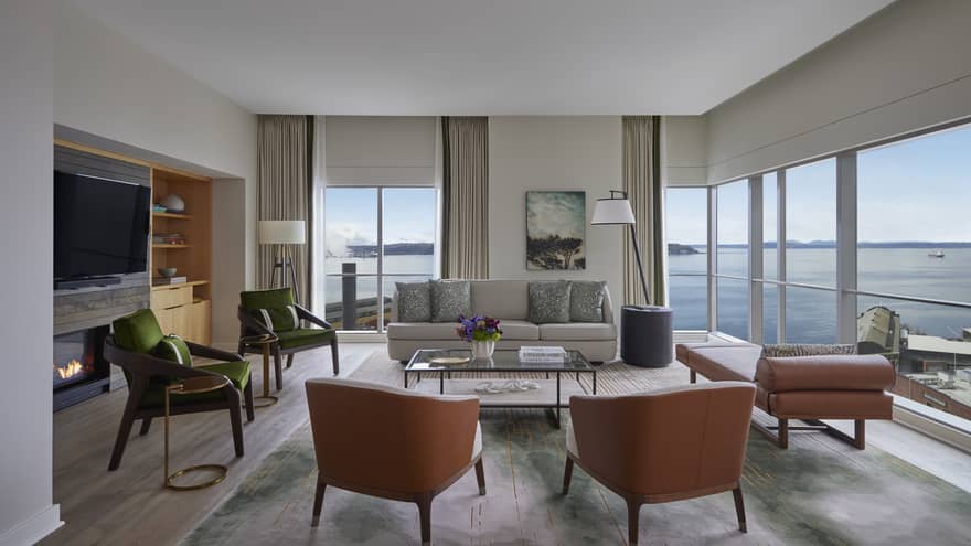 Living room with sofa, four chairs, fireplace and TV, chaise and wrap-around windows looking out to Seattle seafront