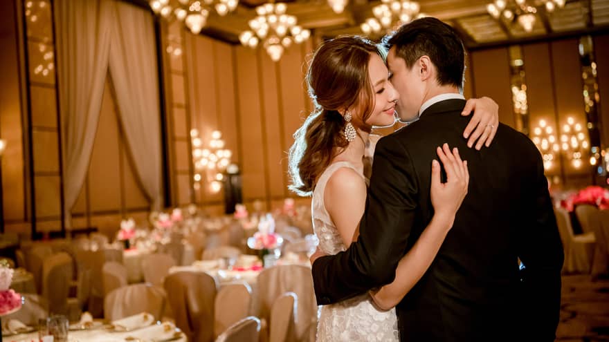A couple embraces while dancing at an elegant wedding reception with chandeliers, warm lighting, and decorated tables, creating a romantic atmosphere