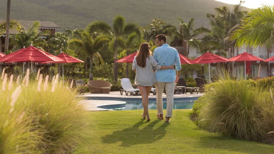 Back of woman with arm around man, strolling across green lawn to outdoor pool