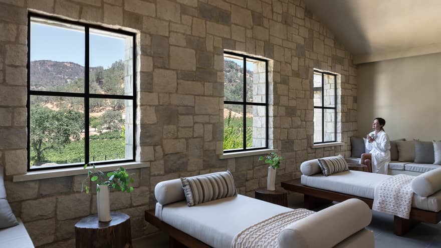Woman in spa robe sits on couch with a tea in hand in large sunny treatment room at Spa Talisa, looking out window at Napa Valley hillside