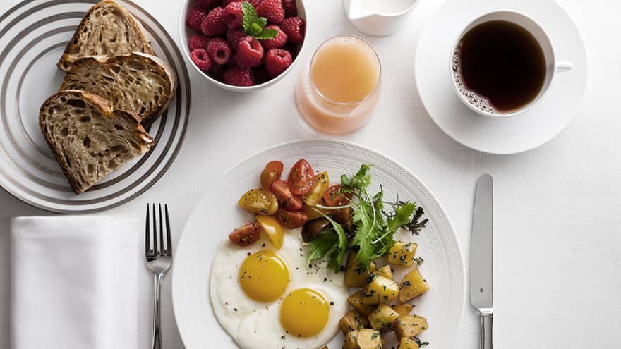 Breakfast plate with sunny-side-up eggs, cherry tomatoes, greens, and roasted potatoes, alongside toast, raspberries, grapefruit juice and coffee.